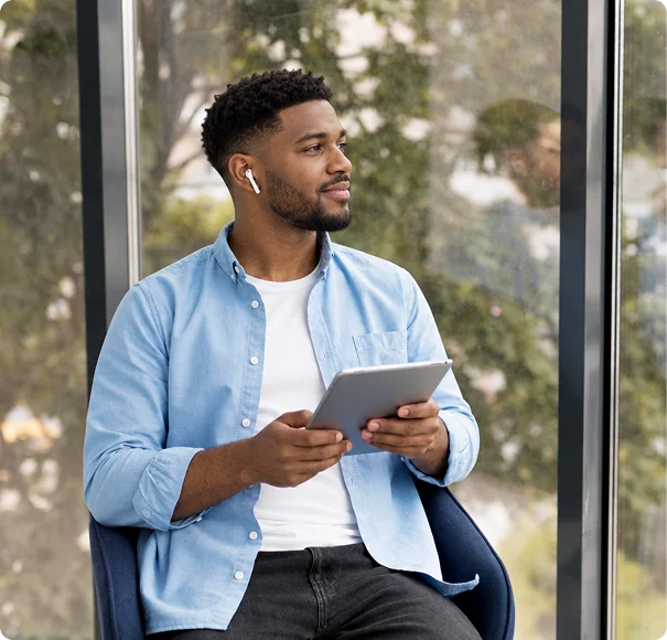Man holding a tablet while sitting indoors near a large window.