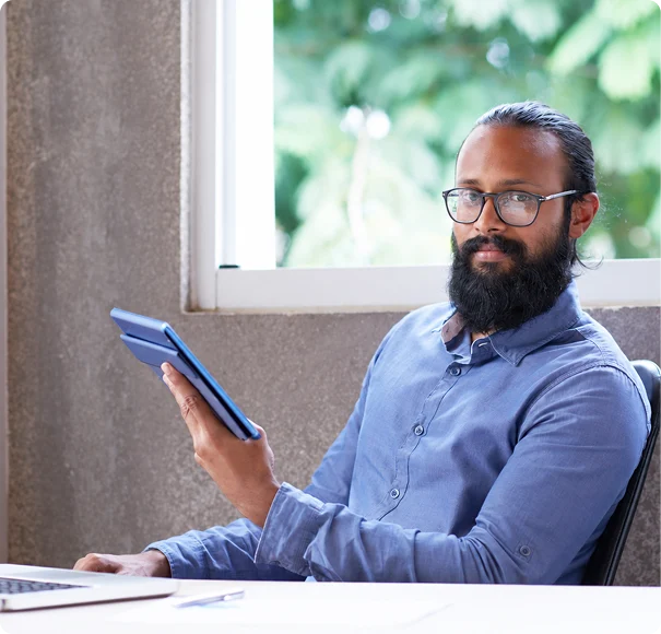 Man holding a tablet while sitting at a desk near a window.